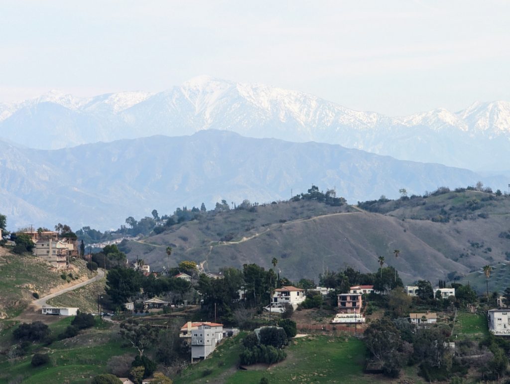 A photo of a snow capped Mt Baldy in the distance. Smaller mountains in the midground and rolling hills of Eastside LA dotted with homes in the foreground.
