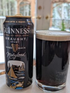 A black can of Guinness Draught next to a glass in which the can's contents have been poured. The liquid in the glass is black with a light tan topper of foam about 3/4" thick. The background is a blurred view of wintry western Maryland woods.