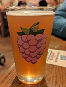 A pint glass of golden amber colored beer. Upon the glass is an illustration of a large boysenberry.