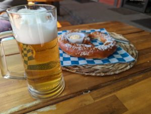 A tall glass of foamy, crisp Kolsch beer next to a large Bavarian style pretzel on a blue and white checkered paper. 