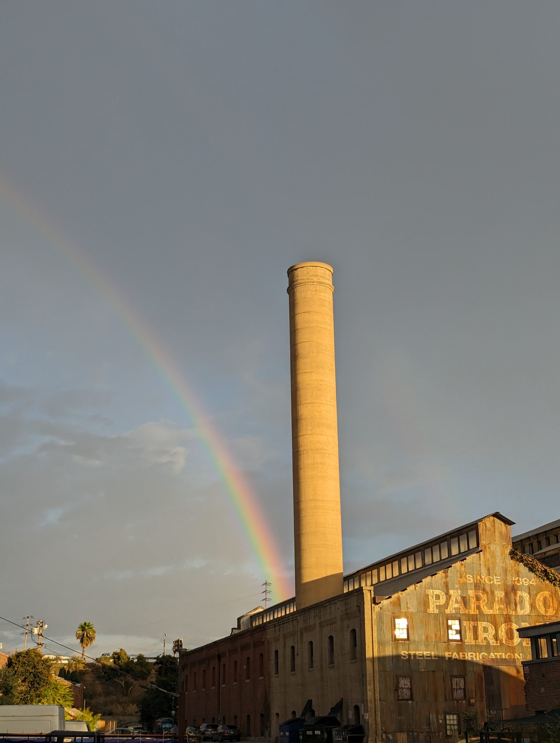 A smoke stack in front of a dark cloudy sky. A rainbow goes from the bottom of the smoke stack off the left of the photo.