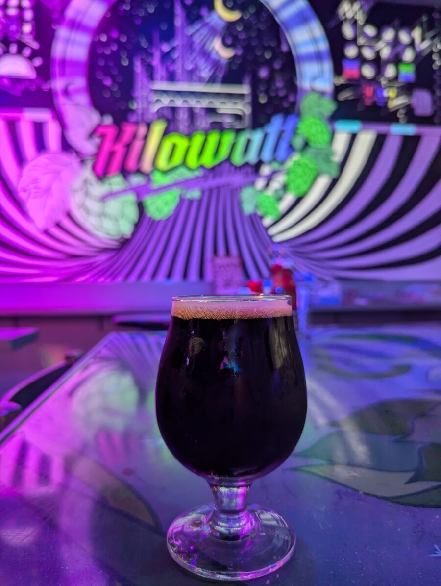 A small glass of a rich and dark Imperial Stout. The background is a colorful mural with the Kilowatt Brewery logo