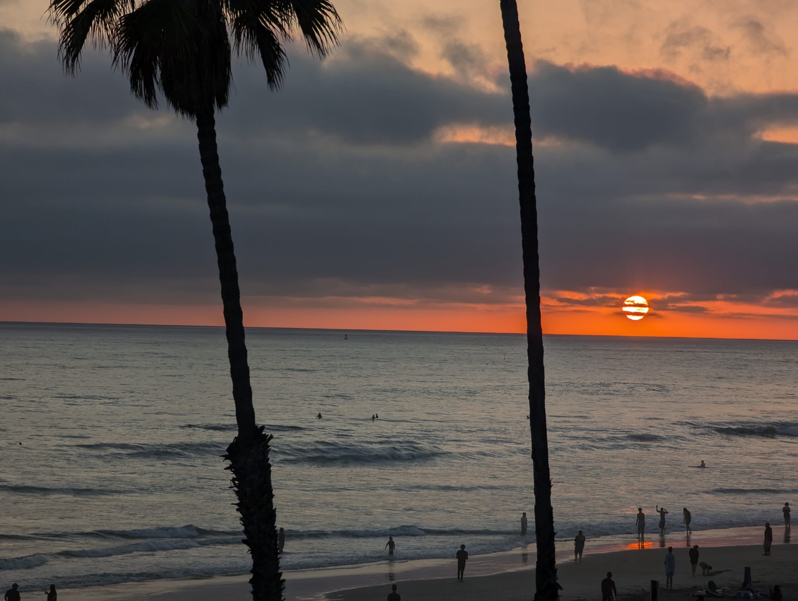 A photo of a red sun setting below the clouds but just above the ocean from the Oceanside pier. Palm trees are silhouetted in the foreground.
