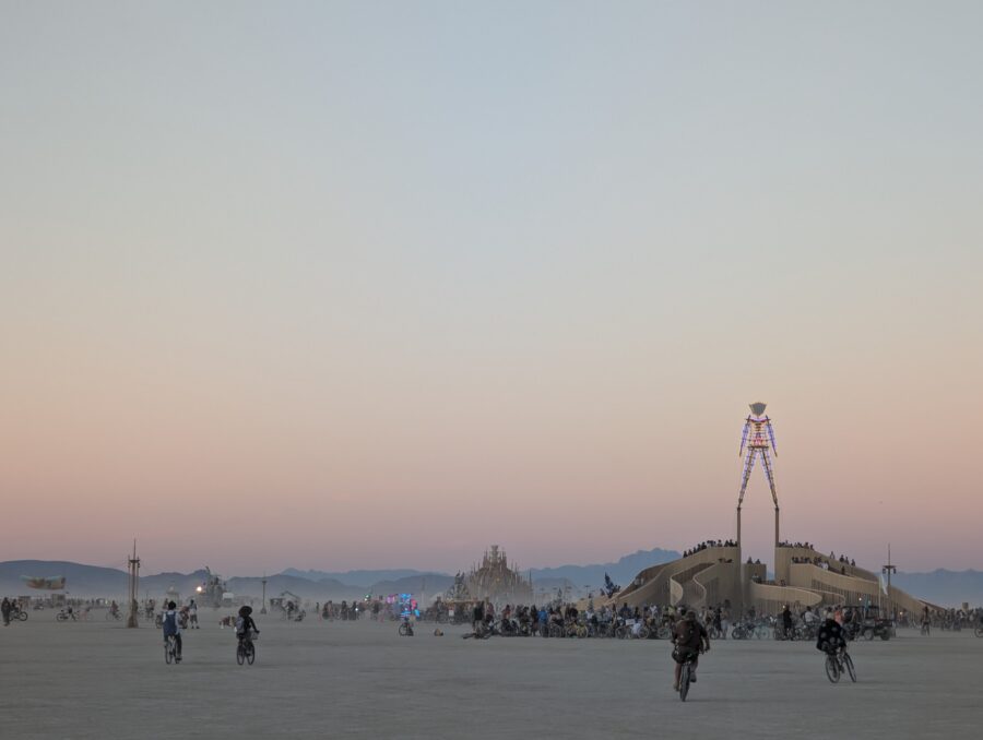 A zoomed in shot of the man standing over his base, the temple to the lower left. It is dusk and the dust is hovering in stillness just above the playa. Citizens ride their bikes in all directions. Some heading home for dinner, others heading out for a night of adventure.