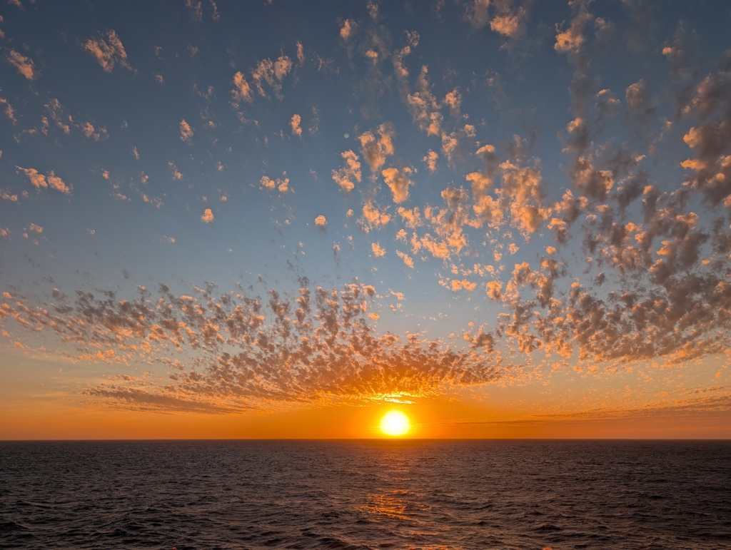 A photo of the sun as it hits the horizon over the Pacific ocean. The clouds dapple the sky and catch the last orange light of the day.