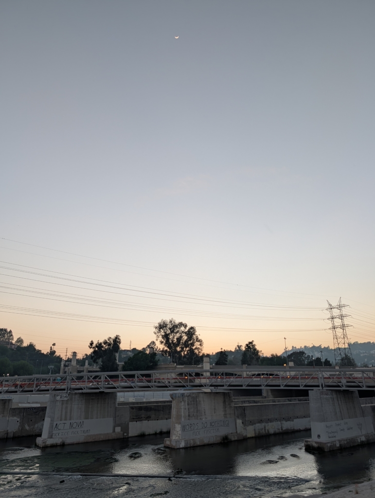 A photo of the Hyperion bridge over the Los Angeles River at sunset. A slim waxing crescent moon hangs in the sky.