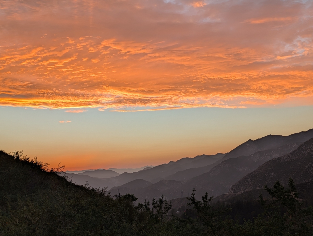 A photo of the mountains in the Angeles National Forest after the sun has set. Bright orange clouds are in the top of the photo with layers of mountains filling the lower part of the photo.